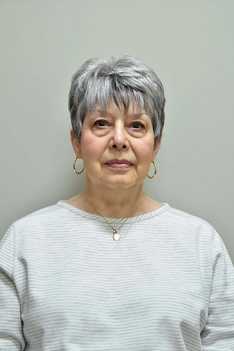 Barbara Bonanno A woman with short gray hair, wearing a striped shirt and gold hoop earrings, stands against a plain background.