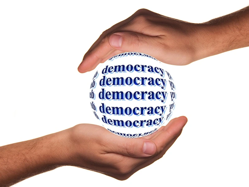 Democracy Image 1 Two hands encircle a sphere covered with the word "democracy" in blue, symbolizing protection or nurturing of democratic values.