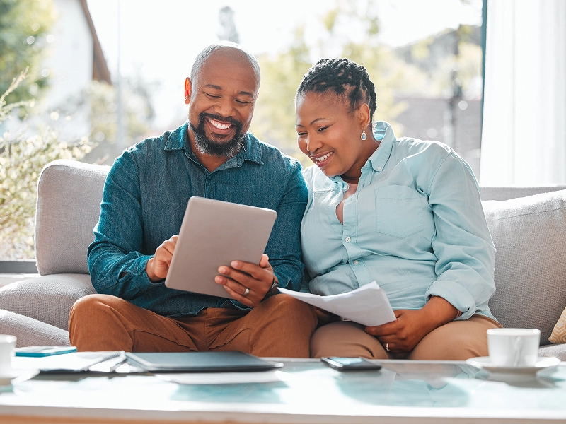 A man and woman, members of the Couple Membership, sit smiling at a tablet on a couch. They hold documents in a well-lit room with a window view.