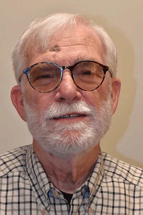 Jerry Henige Elderly man with gray hair and beard wearing glasses and a plaid shirt smiles at the camera.