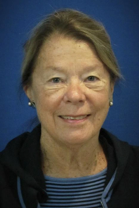 Sharron Nelson A woman with short hair and pearl earrings is smiling, wearing a black jacket over a blue and black striped top, against a blue background.