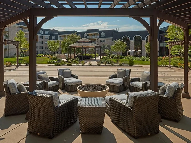 Outdoor seating area with wicker chairs arranged in a circle around a fire pit, under a pergola. A gazebo and spacious garden are visible in the background.