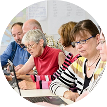 A group of people sit in a row using laptops, focused on their work, in a classroom setting.