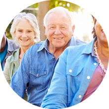 A group of older adults is sitting outdoors, smiling. They're wearing casual clothing, and the background suggests a sunny day.