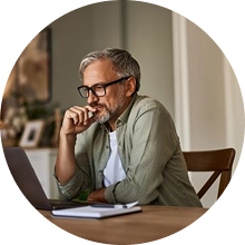 Man with glasses sitting at a table, looking at a laptop screen with a thoughtful expression.
